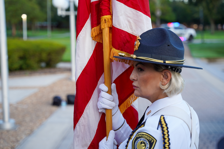 Police officer in uniform holding an American flag during a ceremony, with a patrol car blurred in the background.