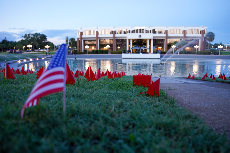 American flags planted in the grass by a pond with a fountain, sunset glow and a lit building in the background.