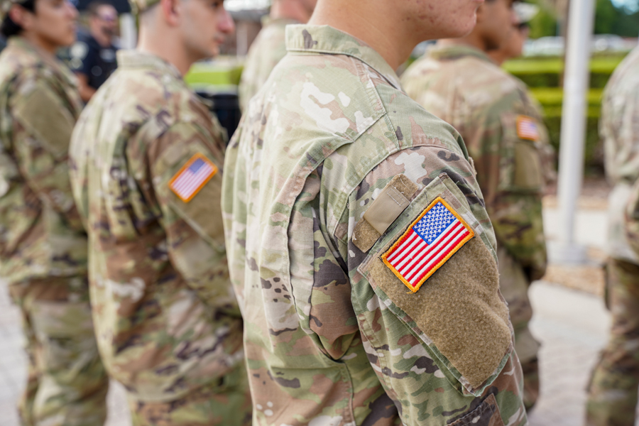 Close-up of a soldier's shoulder in camouflage with a U.S. flag patch on the sleeve, others blurred in the background.