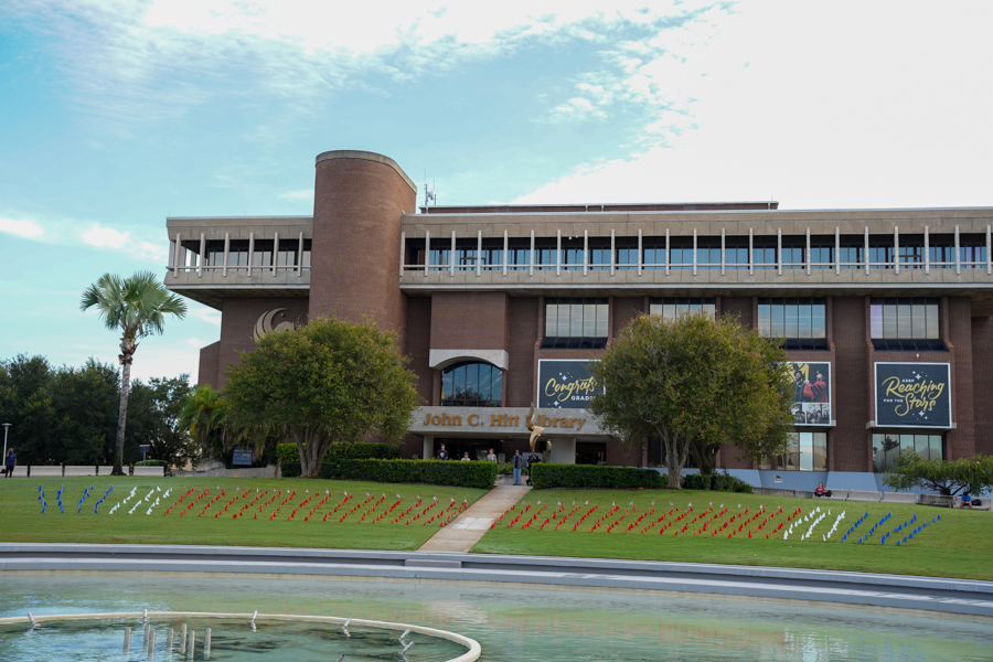 John C. Hitt Library building with a landscaped lawn in front, rows of colorful chairs arranged for an outdoor display, and a circular fountain in the foreground under a blue sky.