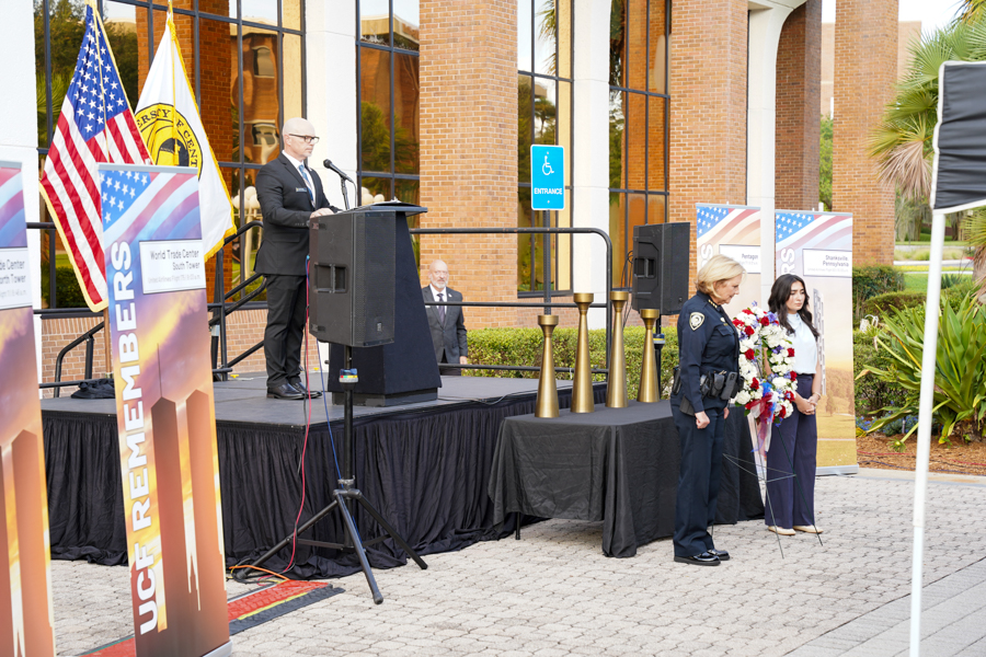 Speaker at a podium addressing an outdoor ceremony, with American flag, banners, wreaths, and uniformed officer nearby.