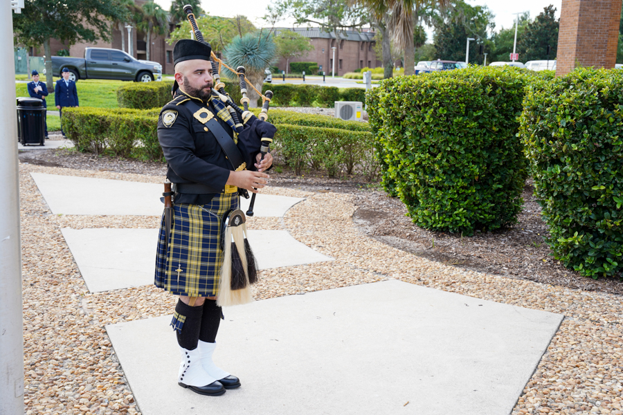 A man in a black jacket and blue plaid kilt plays bagpipes on a gravel path outdoors, with trimmed hedges nearby.