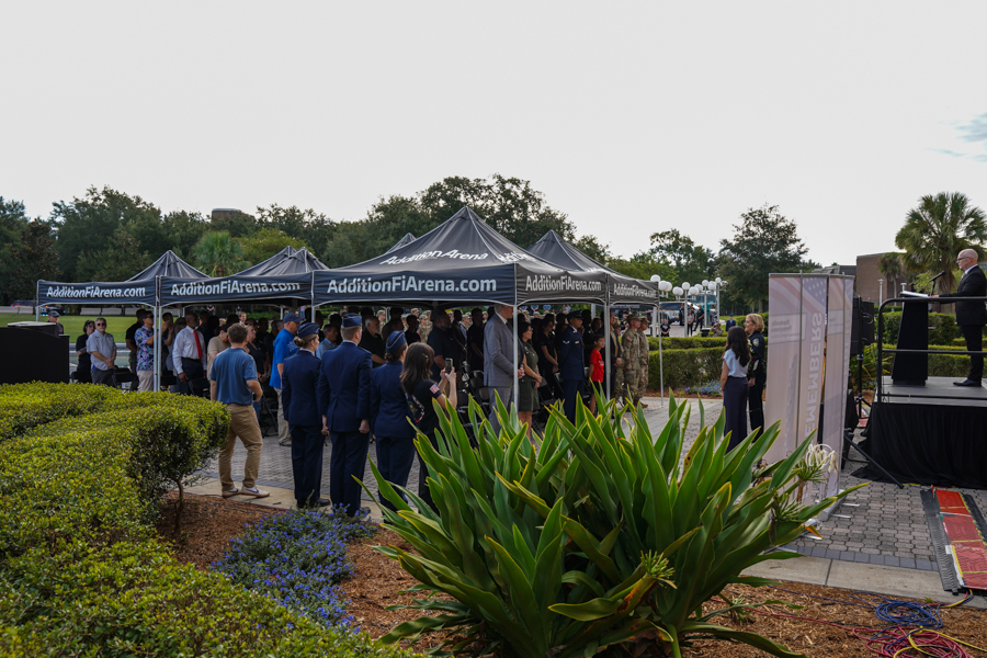 People gathered under black tents with AdditionFiArena.com banners at an outdoor ceremony, crowds facing a stage with a speaker.