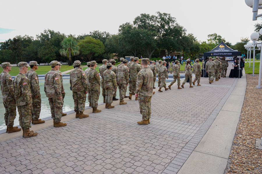 Soldiers in camouflage uniforms lined up along a brick path beside a water feature, with a dark tent and people in the background.