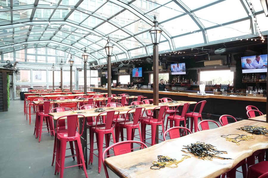Bright industrial-style bar and dining area with red metal chairs and long wooden tables under a glass ceiling.