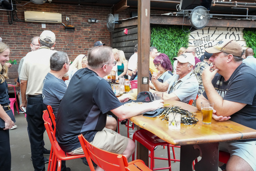 Group of people sitting at a wooden bar table outdoors, drinks in hand, chatting in a brewery setting.]