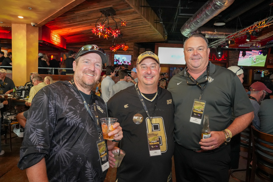 Three men in a crowded bar pose for a photo, each wearing event lanyards and holding drinks, smiling at the camera.