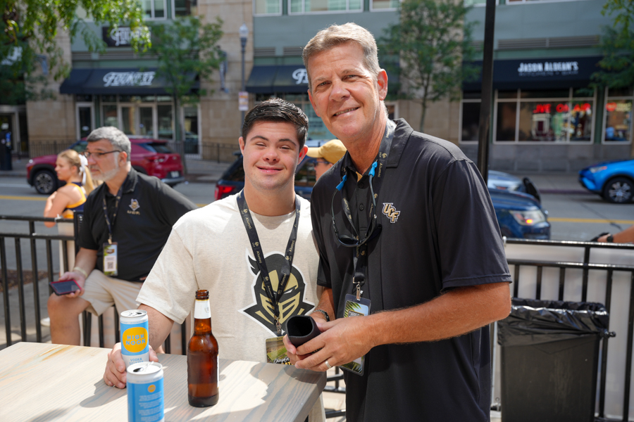 Two men pose for a photo at an outdoor street-side event, drinks on a table in front of them.