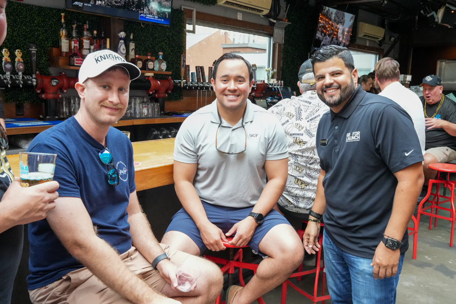 Three men sit and stand around a bar, smiling for the camera in a casual social setting with drinks visible on the counter behind them.