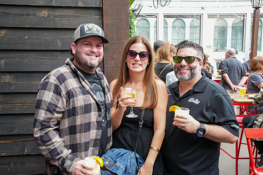 Three adults smiling and holding cocktails at an outdoor event, with a dark wooden wall on the left and a crowd in the background.