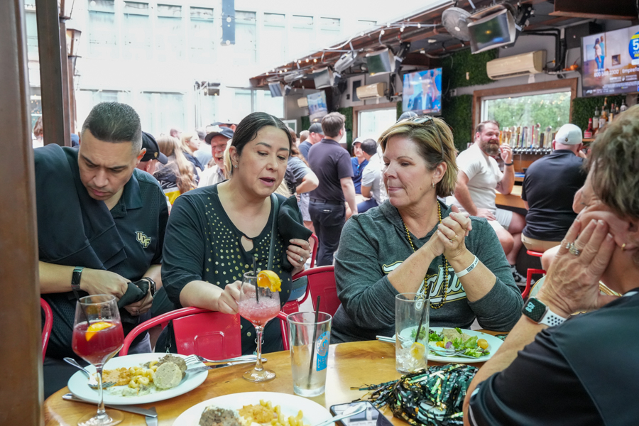 A diverse group sits around a wooden table at a lively bar, enjoying drinks and plates of food while others chat in the background.