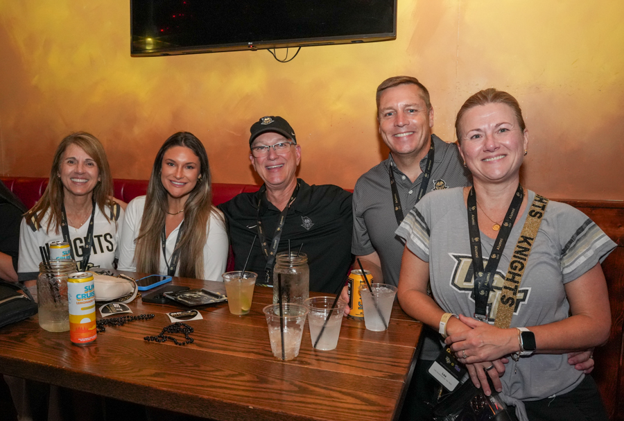 Five adults sit side by side at a wooden table in a bar, smiling at the camera; drinks and event lanyards on the table.