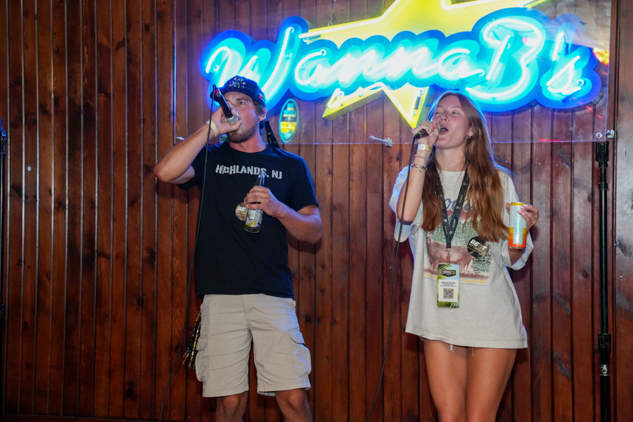 Two people singing into wireless mics on a wooden stage, man holding a beer bottle while the woman holds a cup, with a blue neon sign behind them.