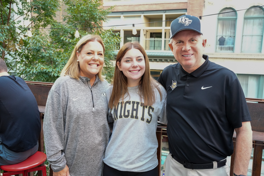 Three people posing outdoors on a balcony, smiling at the camera with trees and string lights in the background