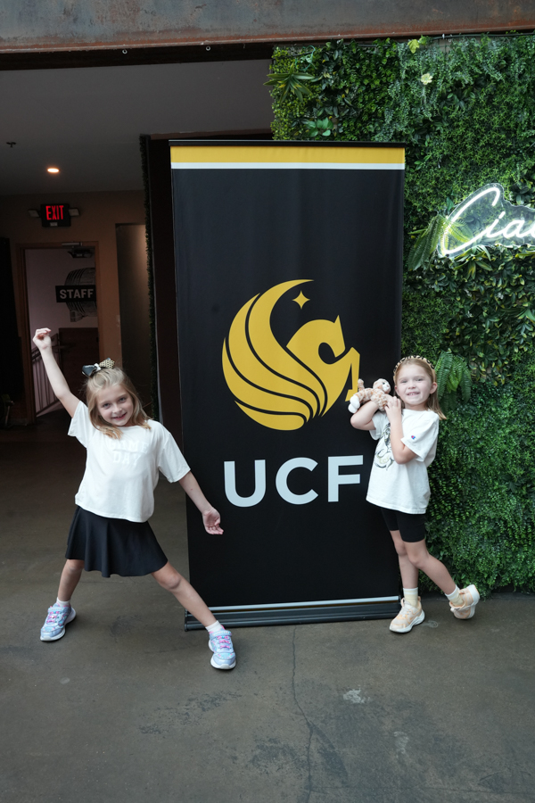 Two smiling young girls pose beside a tall black UCF banner with a yellow logo, one holding a stuffed animal.
