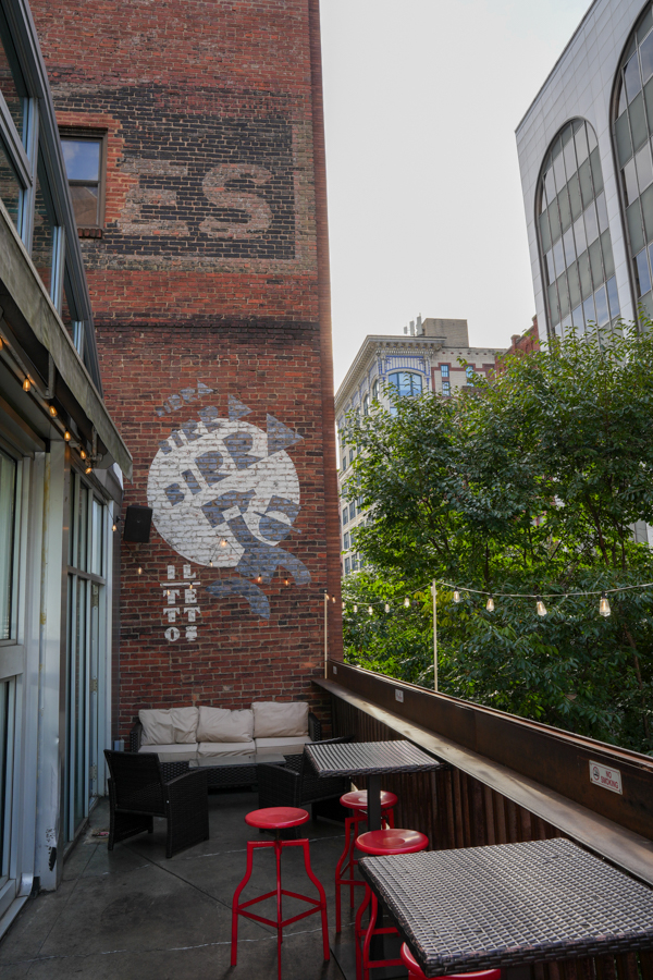 Outdoor city patio with wicker seating, beige cushions, and red stools beneath string lights against a brick wall with faded mural and a circular graphic.