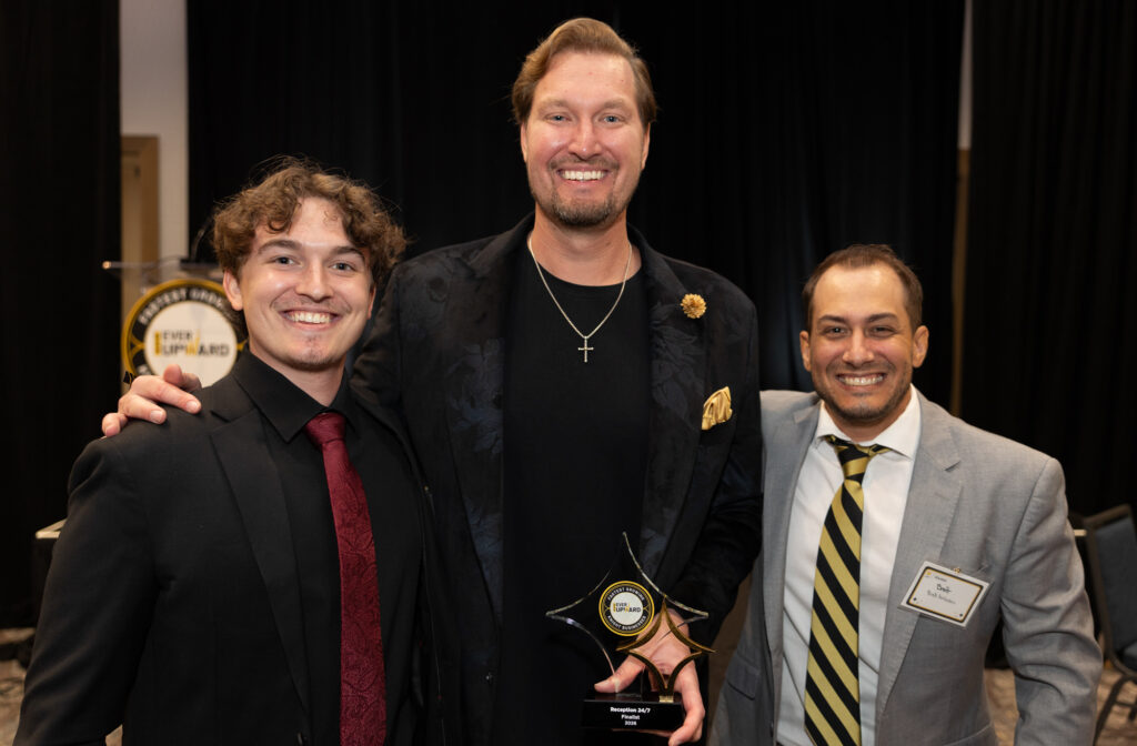 Three men in suits smiling for a group photo at an awards event; the man in the center holds a glass trophy.
