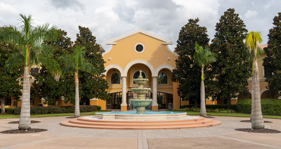 Yellow Mediterranean-style building with an arched colonnade and a multi-tier fountain, surrounded by palm trees in a courtyard.