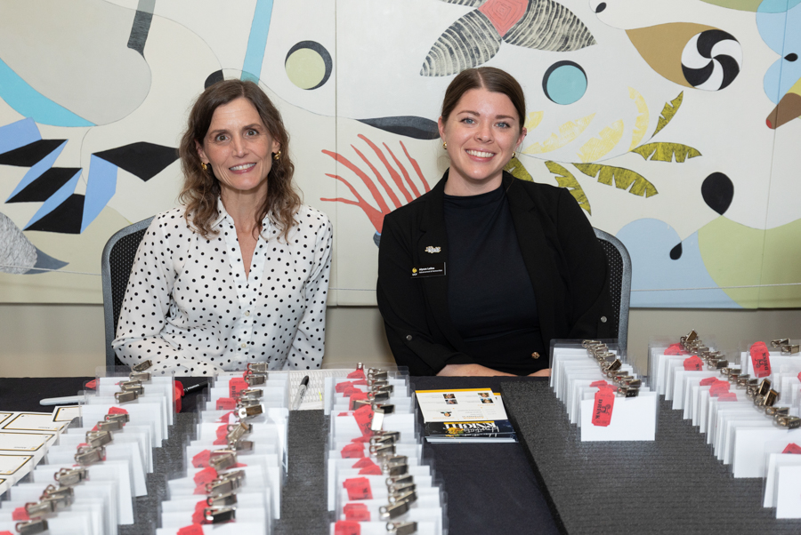 Two women sit at a registration table with rows of name badge holders and lanyards, smiling at the camera.