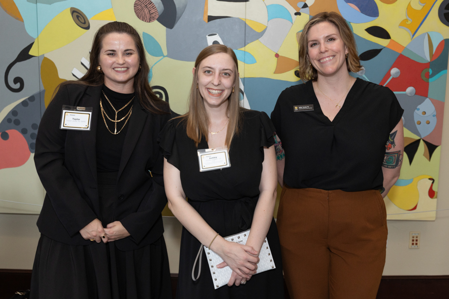 Three women in black outfits posing for a photo at a formal event, all with name badges, with a colorful mural behind them.