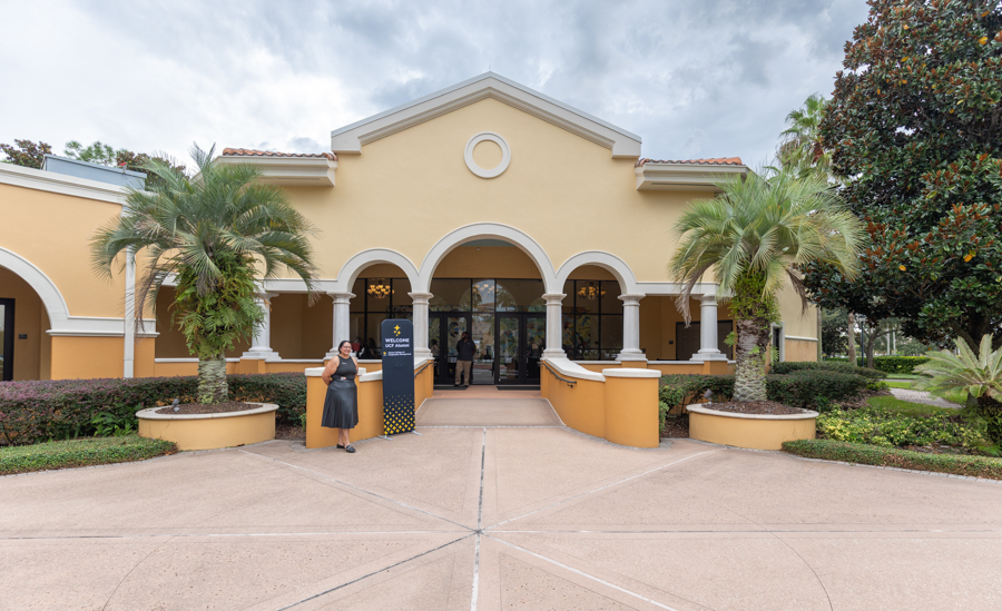 Front view of a yellow building with three arched entrances and palm trees; a woman stands near a welcome sign at the entrance.