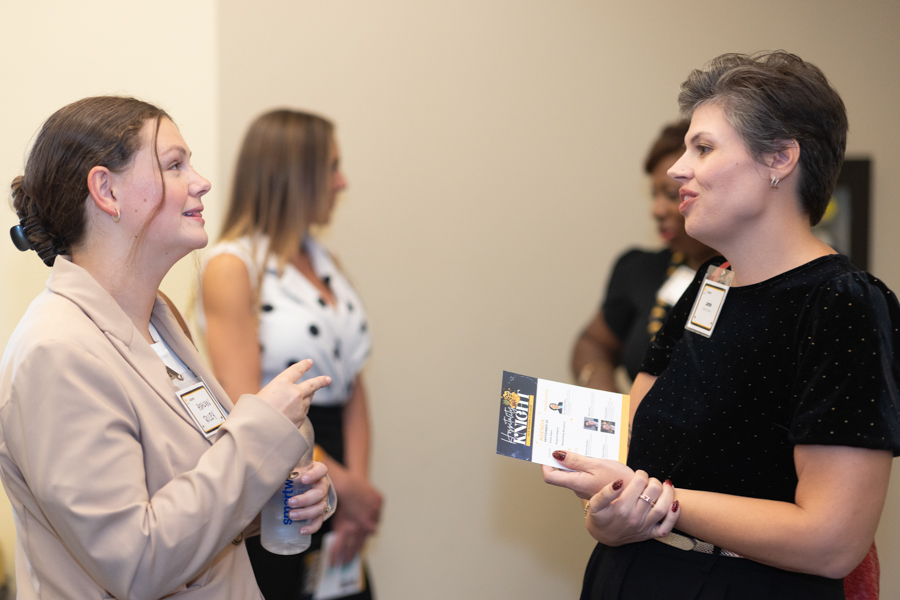 Two women in a hallway wearing name badges converse; one holds a brochure pamphlet-to-hand.