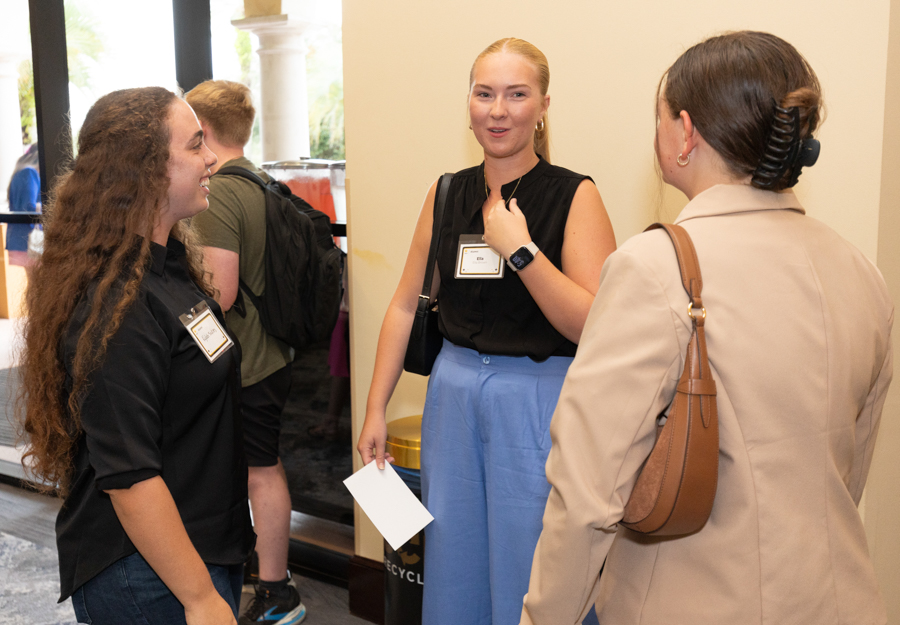 Three people converse at an event: two women with name badges chat with a woman in a beige coat carrying a brown bag near a doorway.