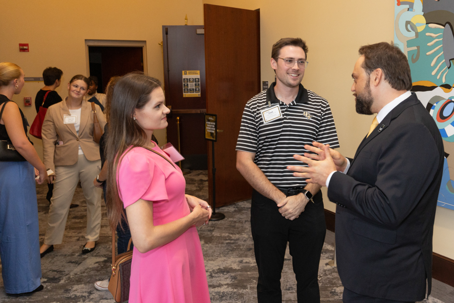 Woman in a pink dress chats with two men in business attire at a networking event in a hallway, name badges visible on all.