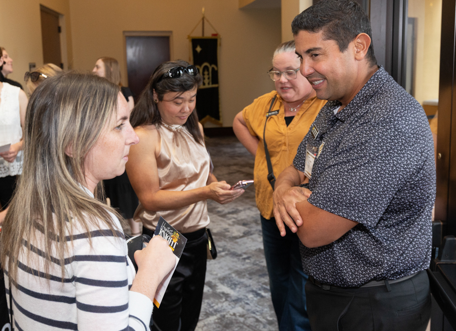A group of adults in a hallway networking; a smiling man in a patterned shirt chats with attendees while two women, one with a striped sweater and another in a peach top, listen.