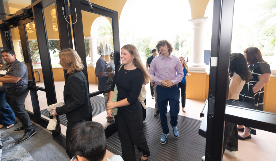 Group of young adults lining up to enter a building through glass doors on a sunny day, with yellow walls and arches in the background.