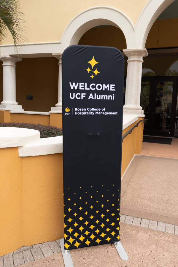 Welcome banner for UCF Alumni outside Rosen College of Hospitality Management with gold star logo on a tall black display
