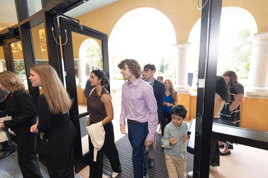 Group of people in smart casual attire walking through large glass doors at an arched, sunlit entrance.
