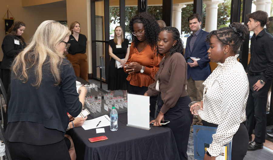 Registration desk at a reception with guests in business attire waiting to check in; a staff member with blonde hair assists them.