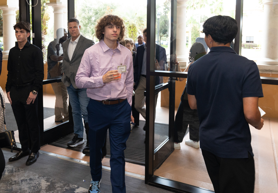 Young man in a lavender shirt and blue pants walks through glass doors, holding a small orange cup and wearing a name tag.