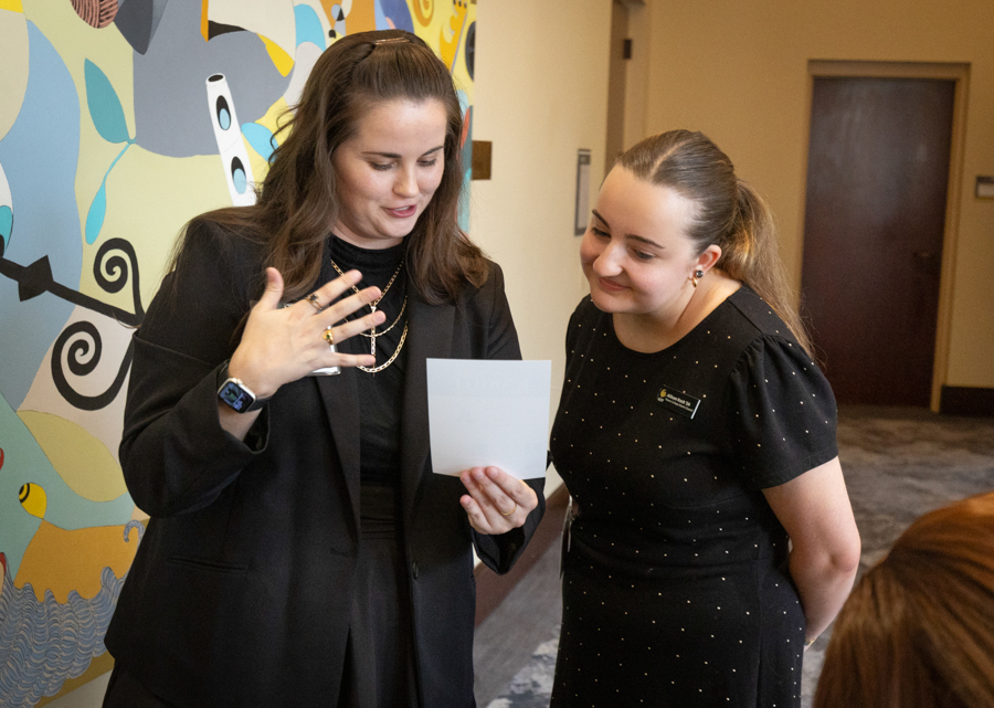 Two women in a hallway, one in a black blazer showing a card to the other in a black dress as they read it together, smiling nearby mural wall