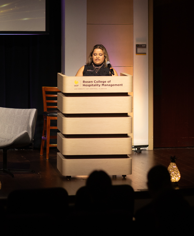 Woman speaks at a podium labeled Rosen College of Hospitality Management, University of Central Florida, addressing an audience.