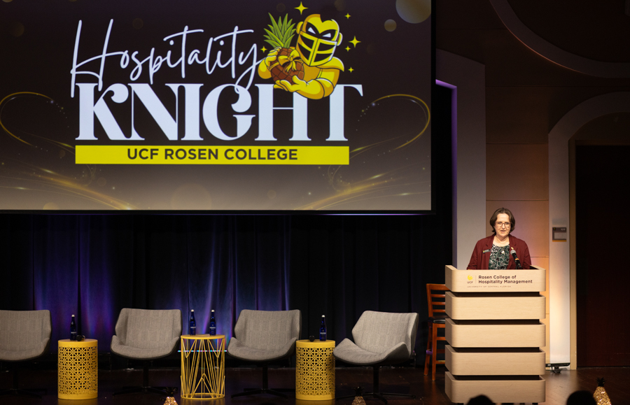 Speaker at a podium on stage during the Hospitality Knight event; screen reads 'Hospitality Knight UCF Rosen College'.