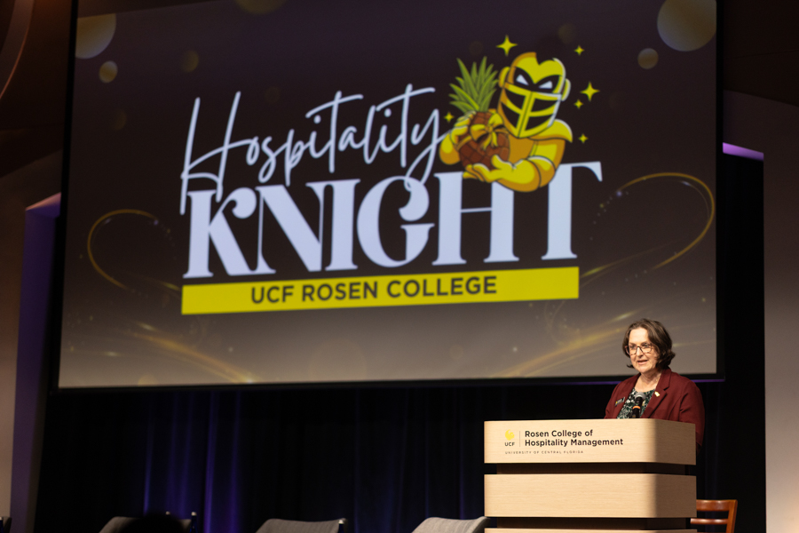 Speaker at a podium during a 'Hospitality Knight' event, with a large screen showing the mascot and 'UCF Rosen College' in the background.