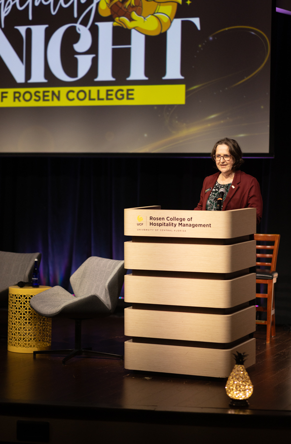 Woman in a maroon blazer speaks at a Rosen College podium on stage, with a large event banner in the background.