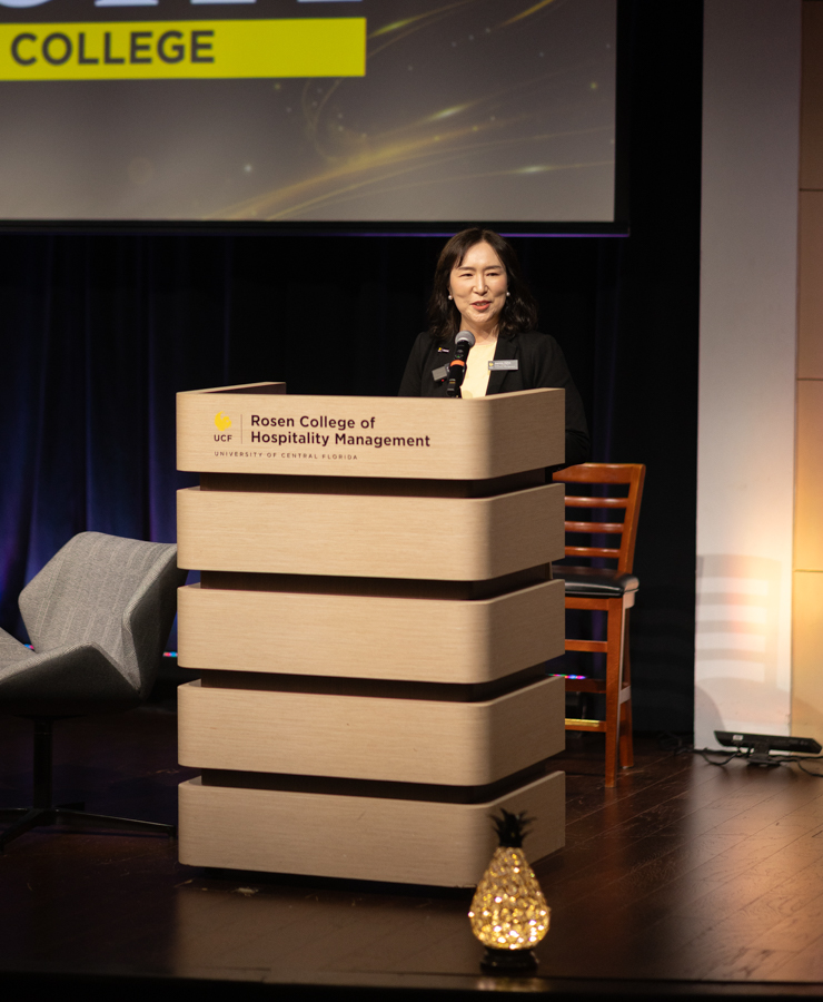 Woman speaking at a podium labeled Rosen College of Hospitality Management on a stage with a dark backdrop and seating nearby.