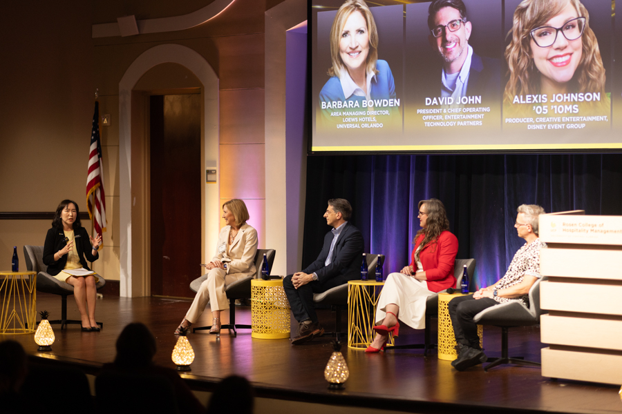Panel discussion on stage with five people seated; moderator speaks from left, audience visible, American flag present, large screen shows bios (Bowden, John, Johnson) above the panel.