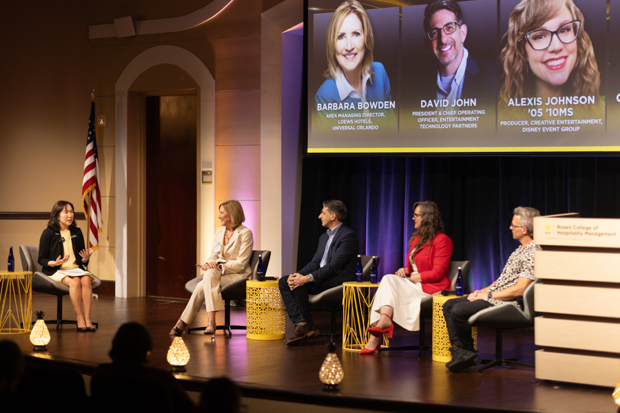 Panel discussion on a lit stage with five speakers seated in a row, moderator at left, American flag nearby and warm decorative lighting.