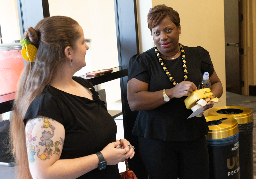 Two women in black clothing converse by a window; one wears a pineapple hair clip and the other holds a yellow purse and water bottle, wearing a beaded necklace.