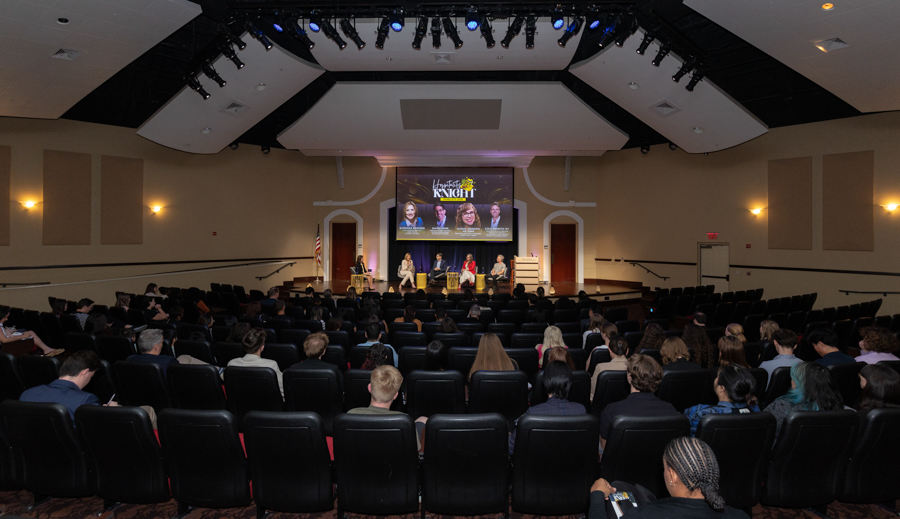 Panel discussion on a stage in a large auditorium with five speakers and a screen behind them showing portraits or slides.