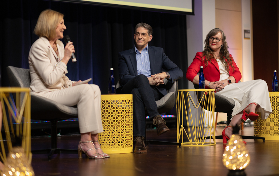 Three panelists on stage: woman in beige suit speaking into a microphone, man in blue blazer, woman in red blazer; yellow tables and water bottles nearby.