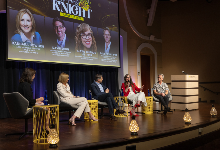 Five panelists seated on a stage in a conference room, with a large screen behind them listing panelists' names and photos.