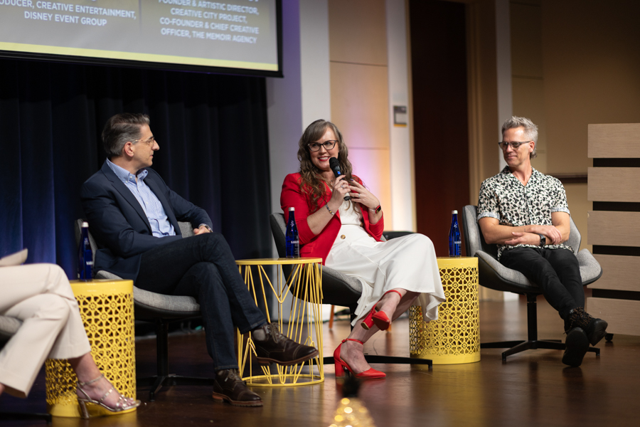 Panel discussion on stage with three speakers; woman in red blazer speaks into a microphone, flanked by two men.