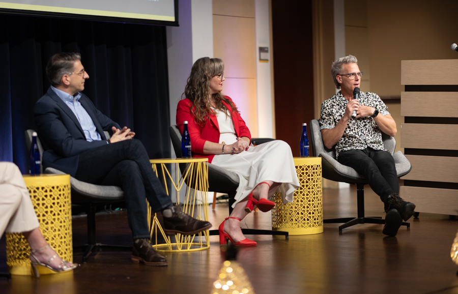 Panel discussion on stage with three people seated in chairs; man in blazer on left, woman in red blazer center, man on right speaks into a microphone.