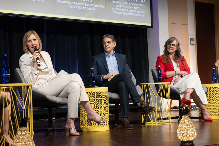 Three panelists on a stage during a discussion: a woman on the left speaks into a microphone, a man in the middle, and a woman in red on the right.