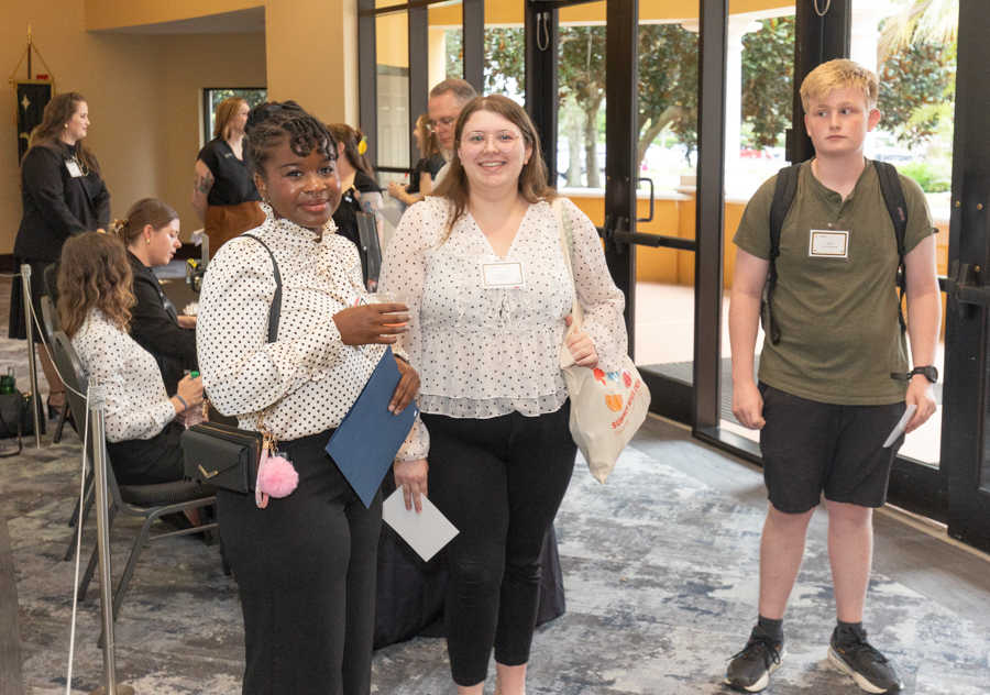 Three attendees at a check-in area smile for the camera: two women in polka-dot blouses in the foreground, a teenage boy with a backpack at the door in the background.
